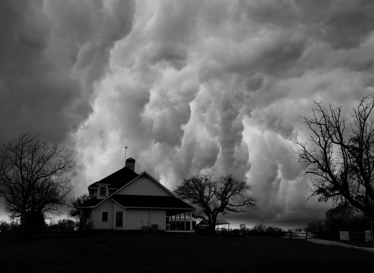 A white farmhouse beneath a towering storm cloud, in black and white