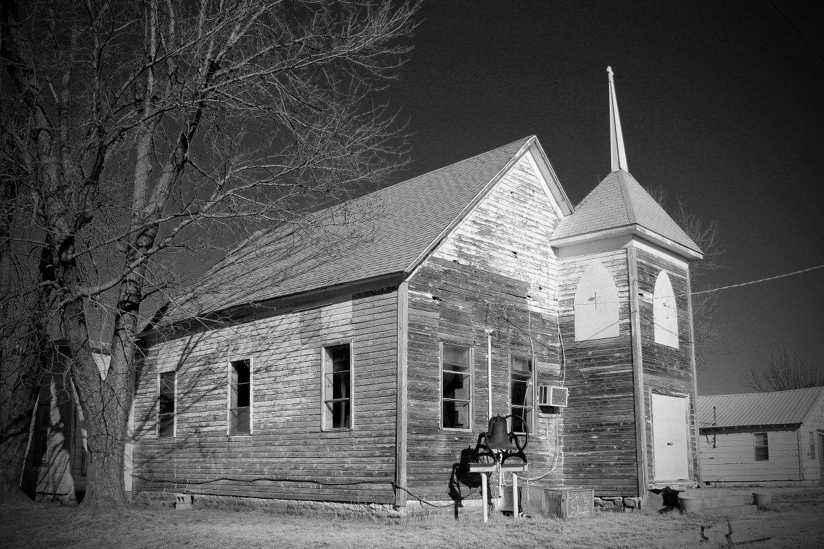 An abandoned clapboard church beside a bare tree, in black and white